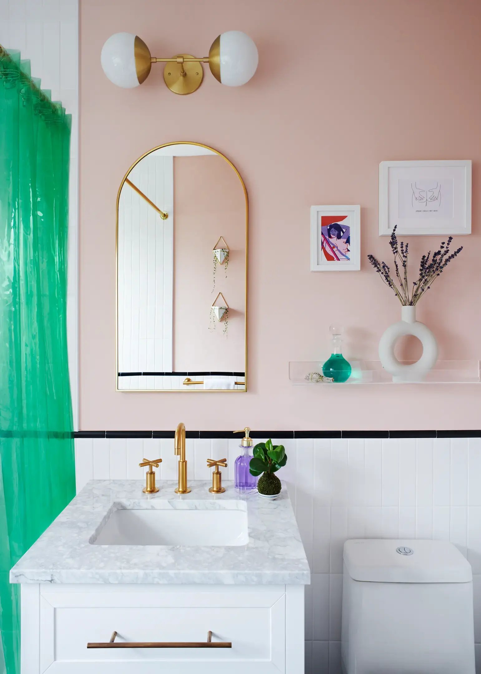 A vanity with a white marble countertop and a white cabinet with gold hardware sits below an arched mirror and a gold double-globe sconce, all against a blush-pink bathroom wall with a green shower curtain visible on the left.