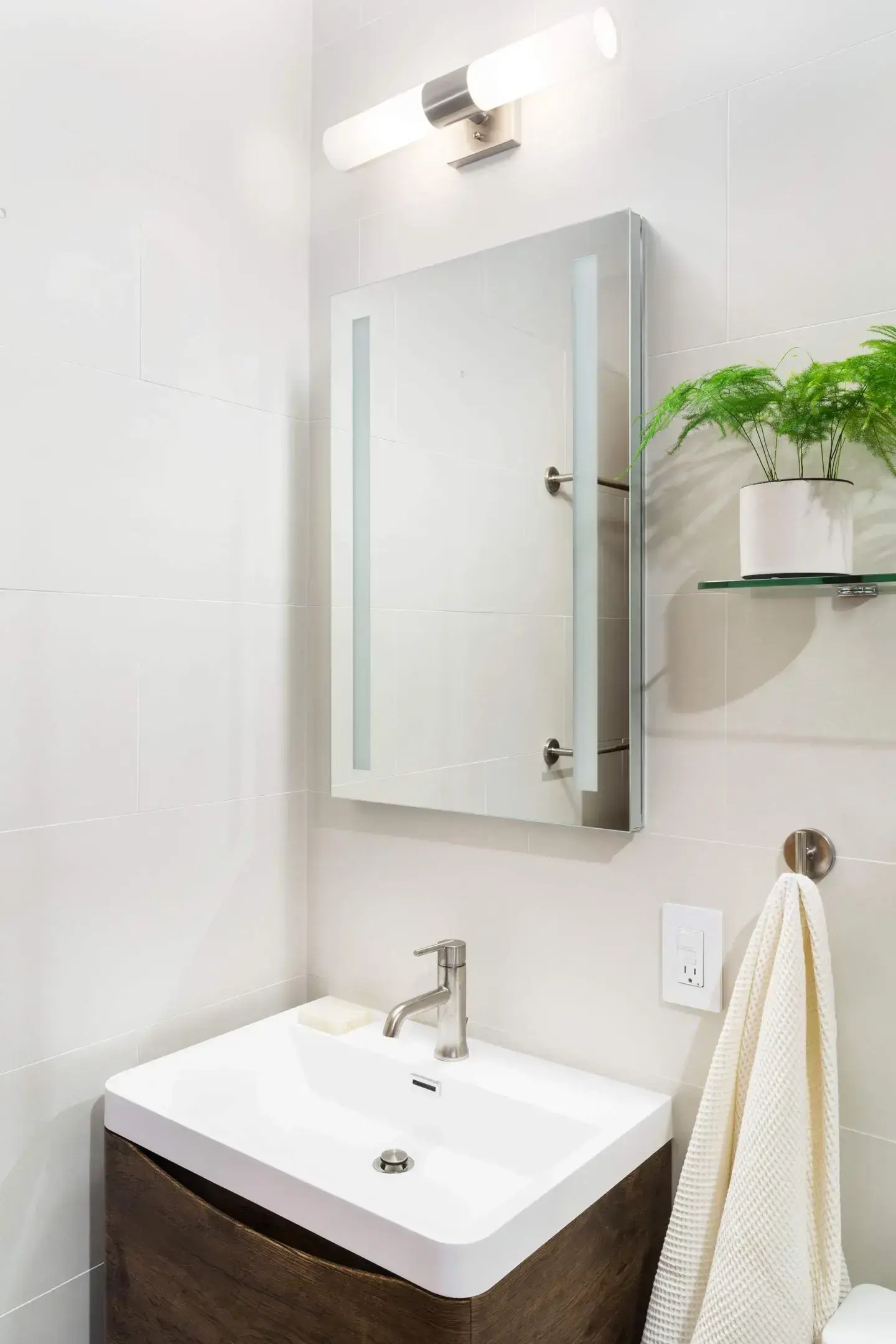 A modern bathroom vanity with a white sink, wood cabinet, lighted mirror, and overhead light fixture.