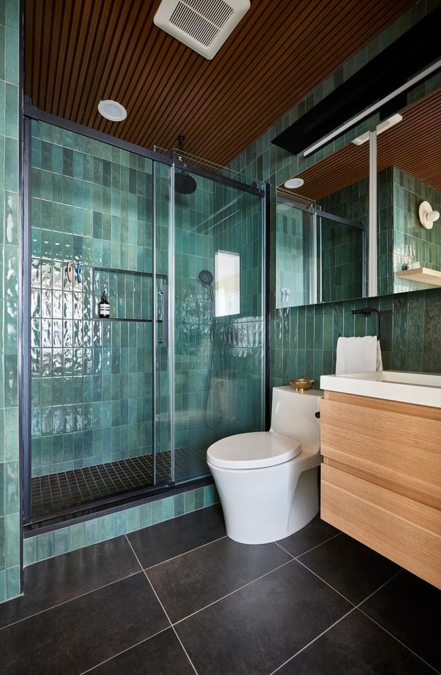 Green tiled bathroom with a wood vanity and slat ceiling.