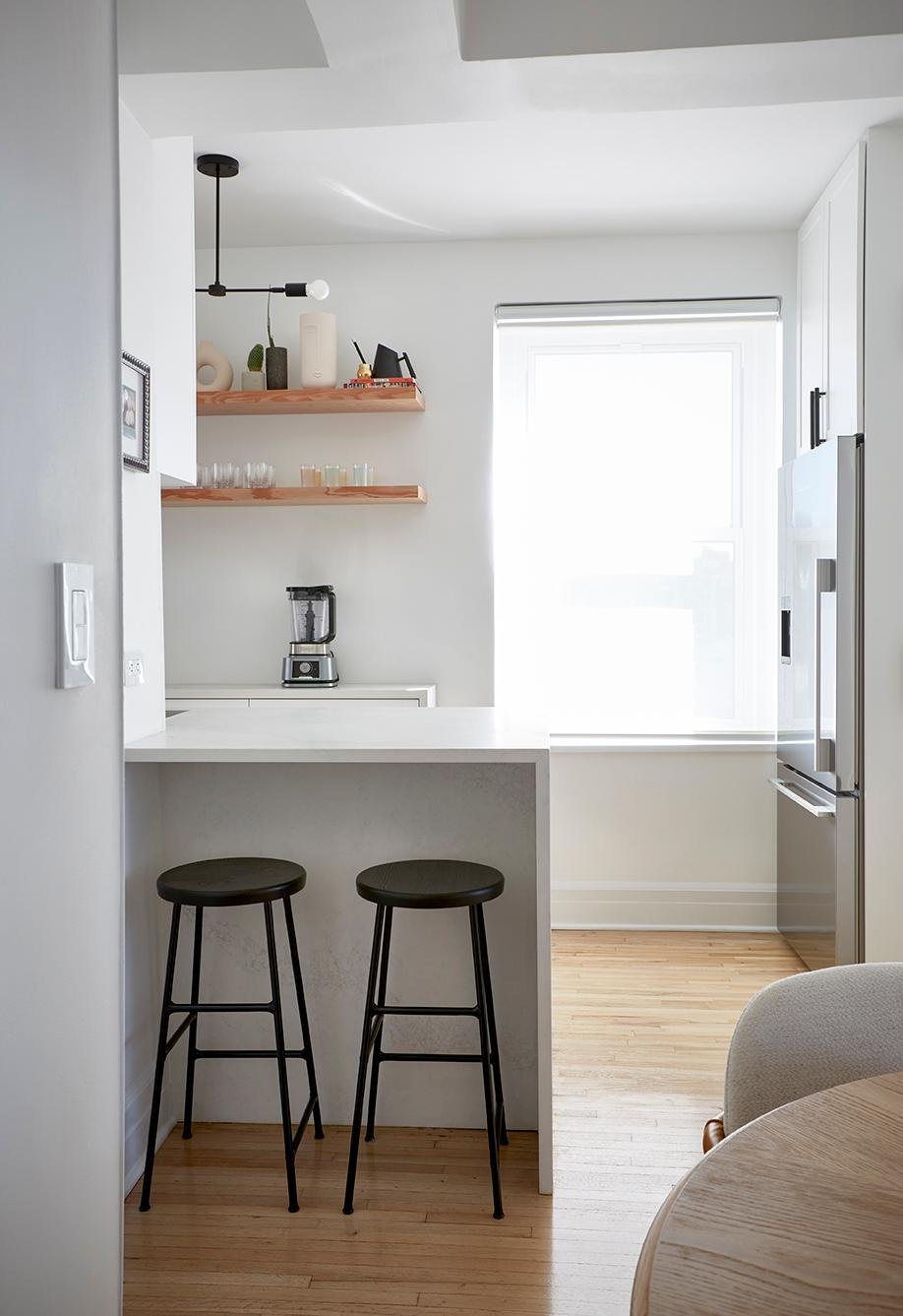 A bright, minimalist kitchen features a white countertop peninsula with two black metal stools, a white base cabinet with a blender, two light-wood open shelves, white upper cabinets, and a tall stainless steel refrigerator next to a large window.