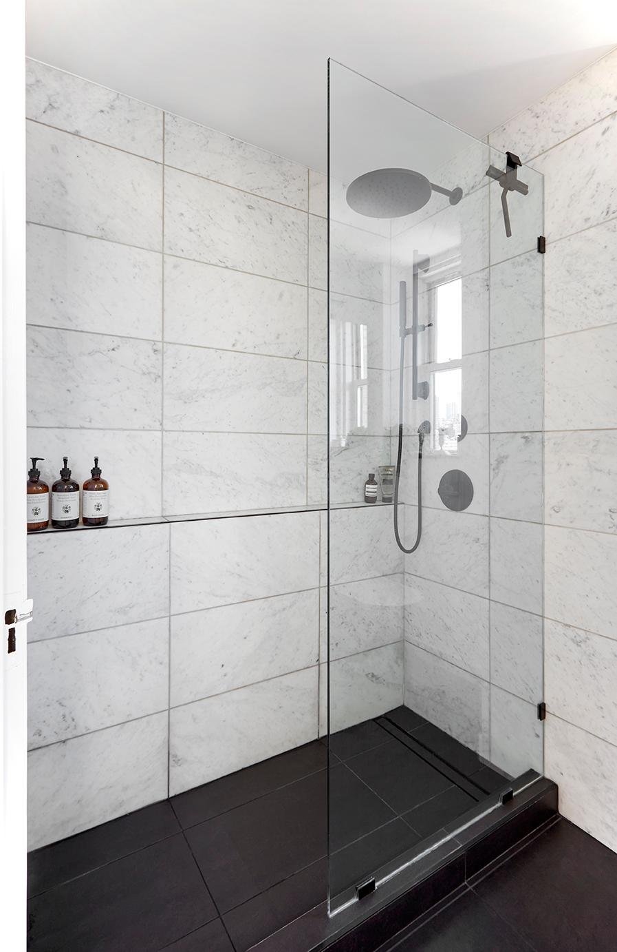 Frameless glass walk-in shower with marble wall tiles, black floor tiles, built-in niche shelving, and a ceiling-mounted rain showerhead for a clean, modern spa-style look.