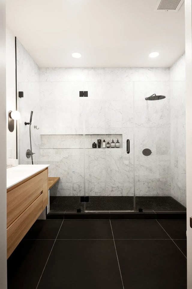 The image depicts a modern bathroom with a glass-enclosed walk-in shower featuring marble tile walls, a built-in shower bench, and a dark tile floor.