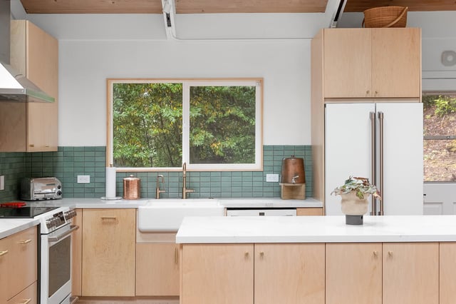 A bright kitchen featuring light wood cabinetry, white countertops, a farmhouse sink, a stainless steel refrigerator, and a backsplash of small, greenish-blue square tiles.