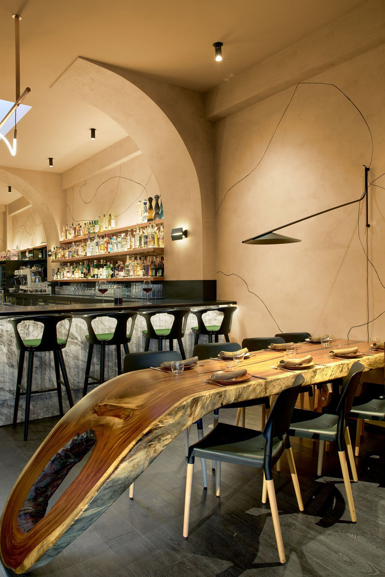 A restaurant interior features a long, live-edge wood dining table with an organic hole near one end, dark chairs, and a bar area with black stools in front of a white marble counter and arched wall niches holding liquor bottles.