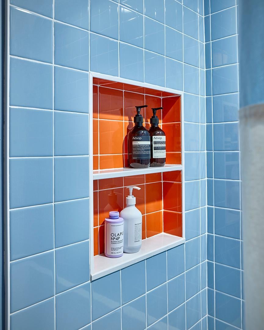 A blue tiled shower wall featuring a recessed orange tiled niche holding bottles of Aesop and Olaplex products.