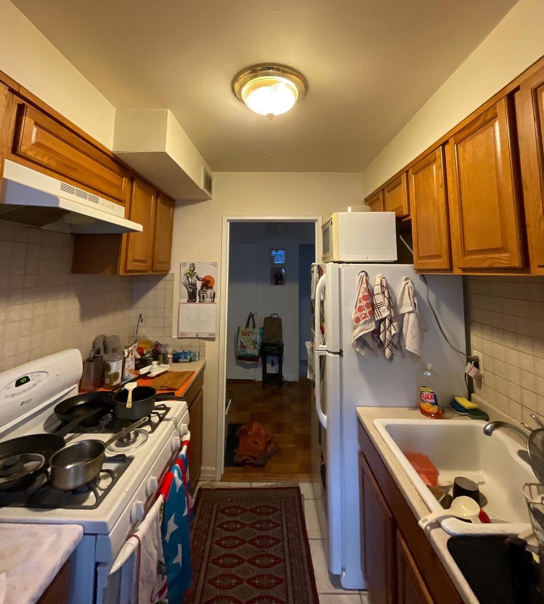 A narrow galley kitchen with wooden cabinets, a white stove and sink, and a refrigerator with towels hanging on it.