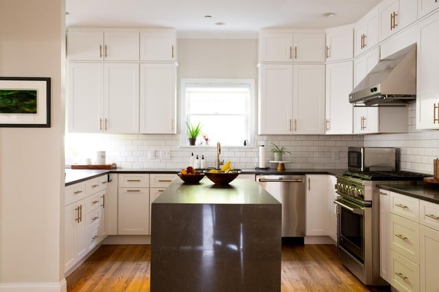 A bright, modern kitchen with white cabinets, gold-colored hardware, a dark countertop on the center island, stainless steel appliances, and wood flooring.