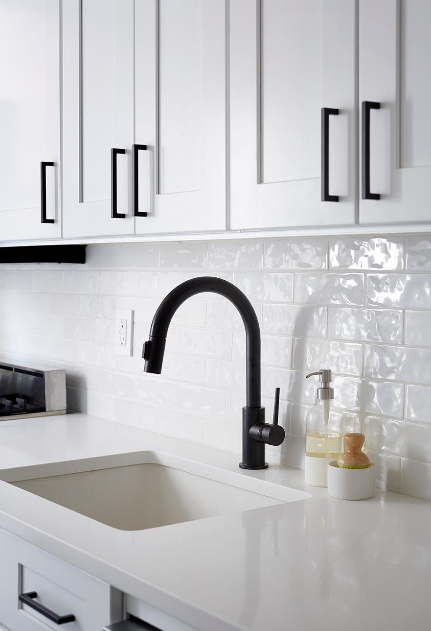 A modern kitchen sink area with white cabinets and countertops, a white subway tile backsplash, and a matte black gooseneck faucet with matching cabinet handles.