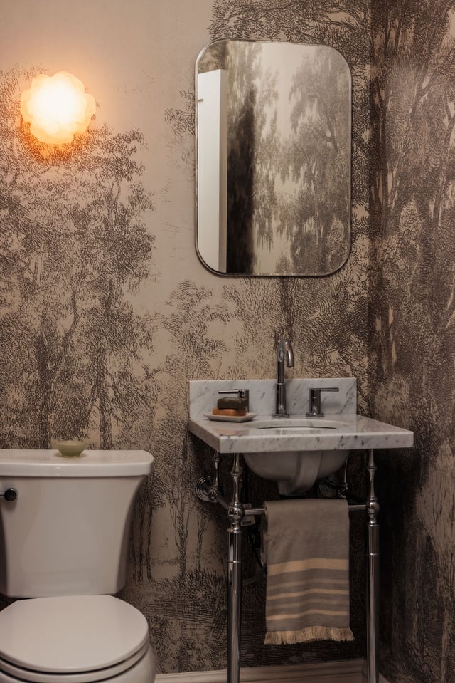 A small bathroom featuring a white toilet, a marble console sink with chrome legs, a rectangular mirror, and monochromatic forest-patterned wallpaper.