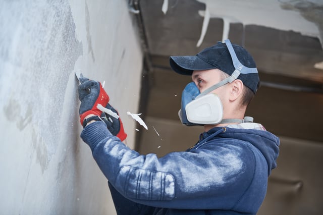 Worker wearing a respirator mask scraping paint off a wall.