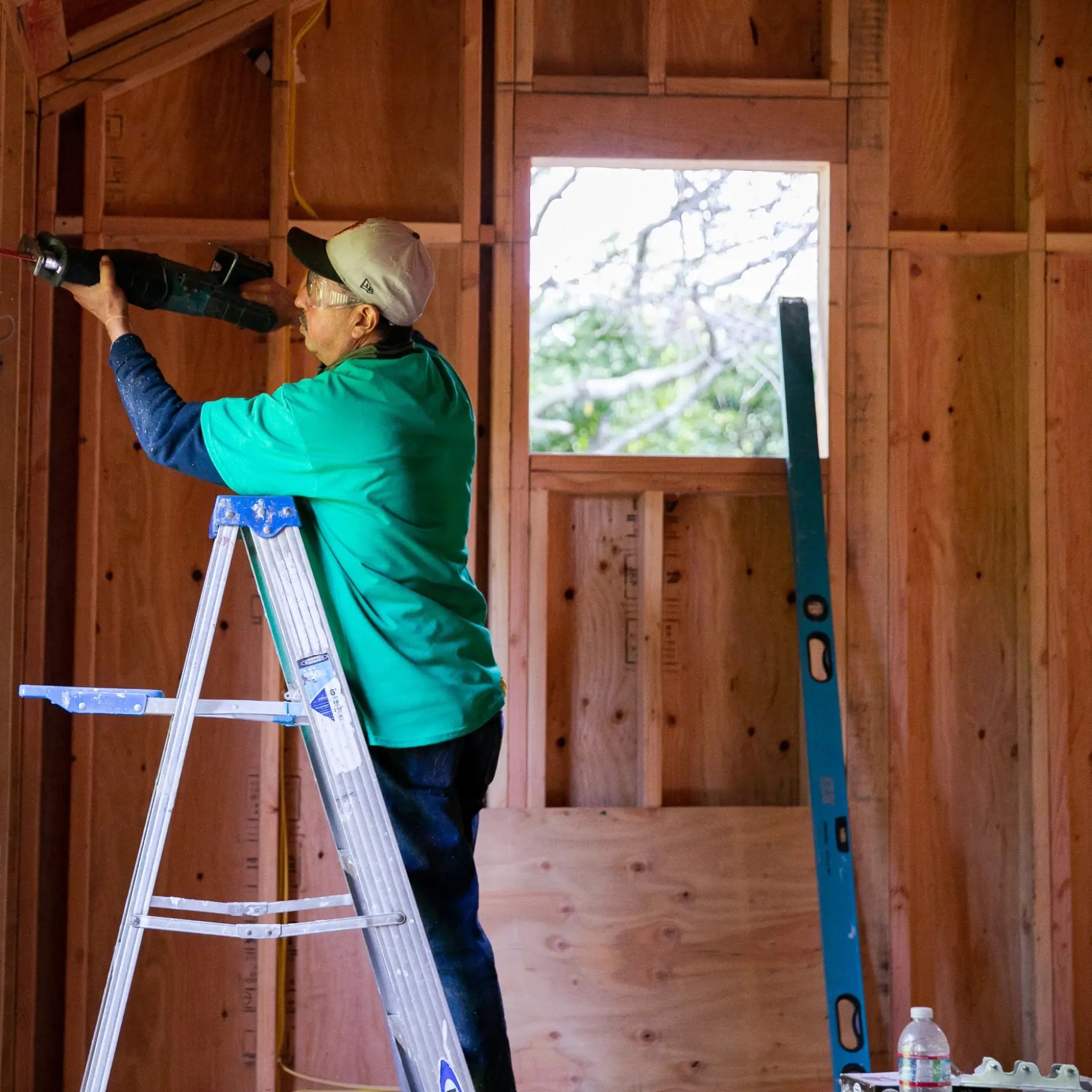 A worker on a ladder sawing wood framing inside an attic.