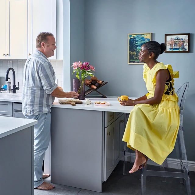 A man and a woman talking in a kitchen with gray cabinets and light blue walls.
