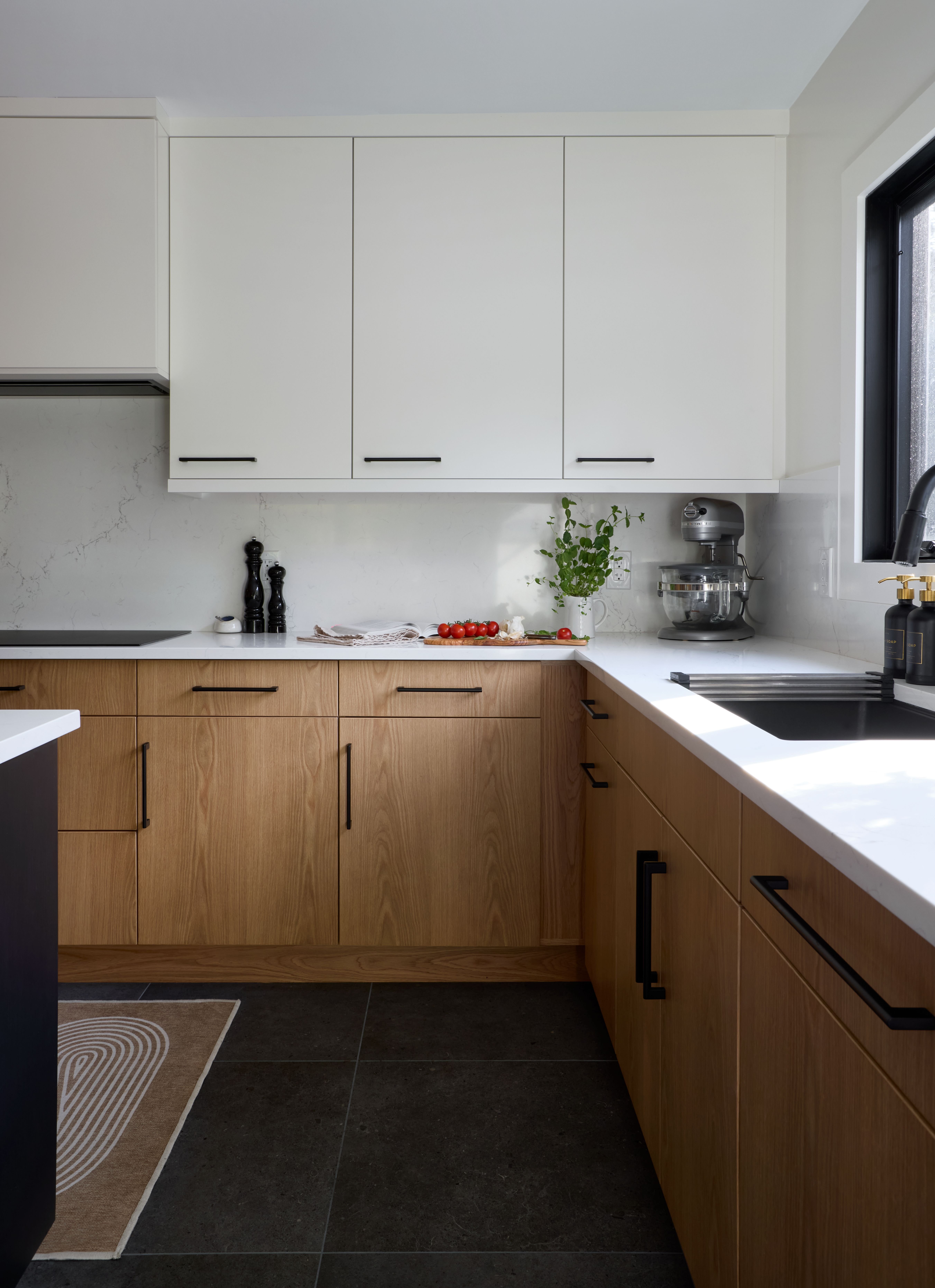 A contemporary kitchen with white upper cabinets, warm wood lower cabinets, white countertops, black hardware, and a clean, minimalist look accented by plants and small appliances.