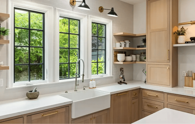 A modern kitchen with light wood cabinets, white countertops, a farmhouse sink, and three large black-framed windows.