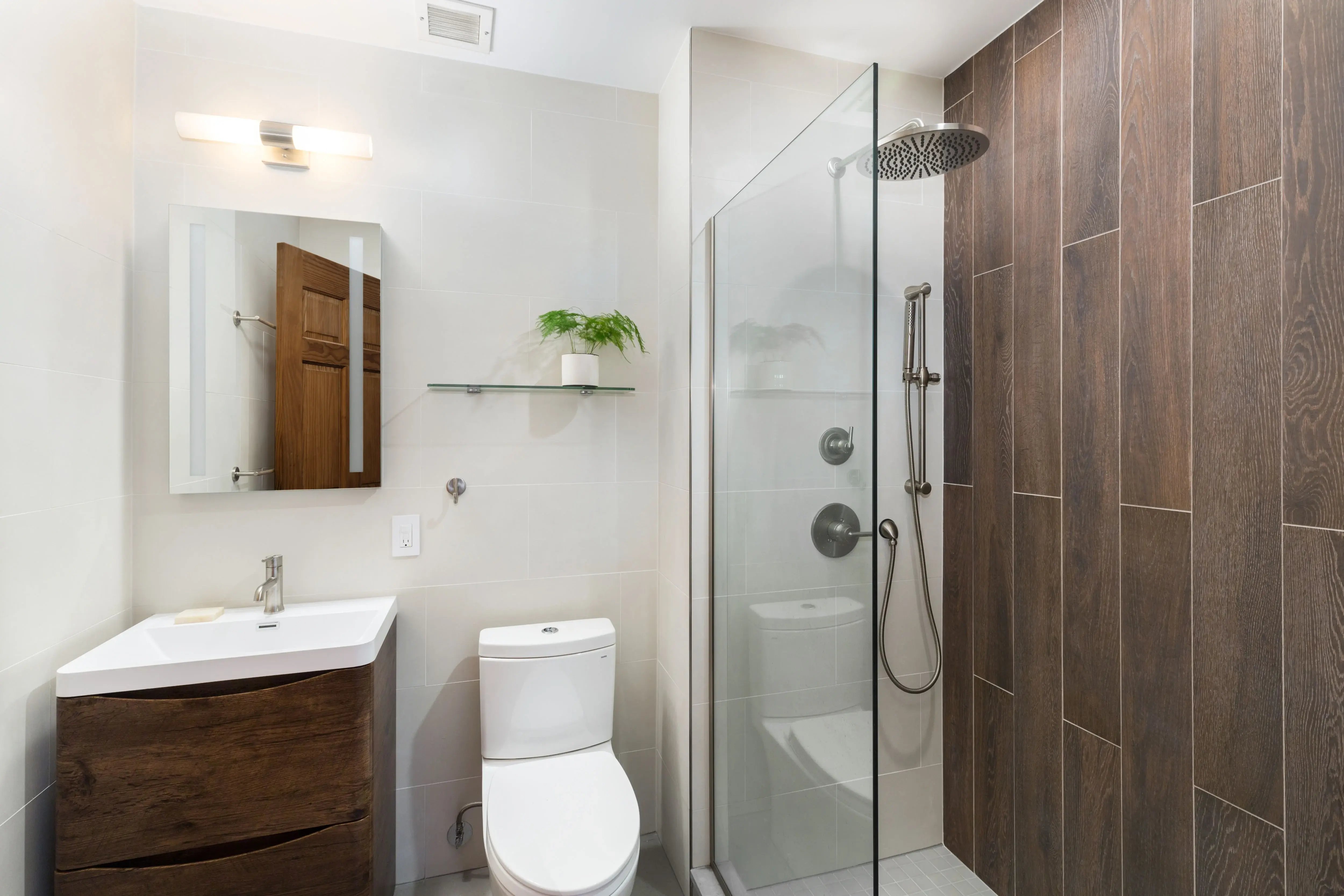 A modern bathroom featuring a floating wood vanity, a white toilet, and a glass-enclosed shower with dark wood-look wall tiling.