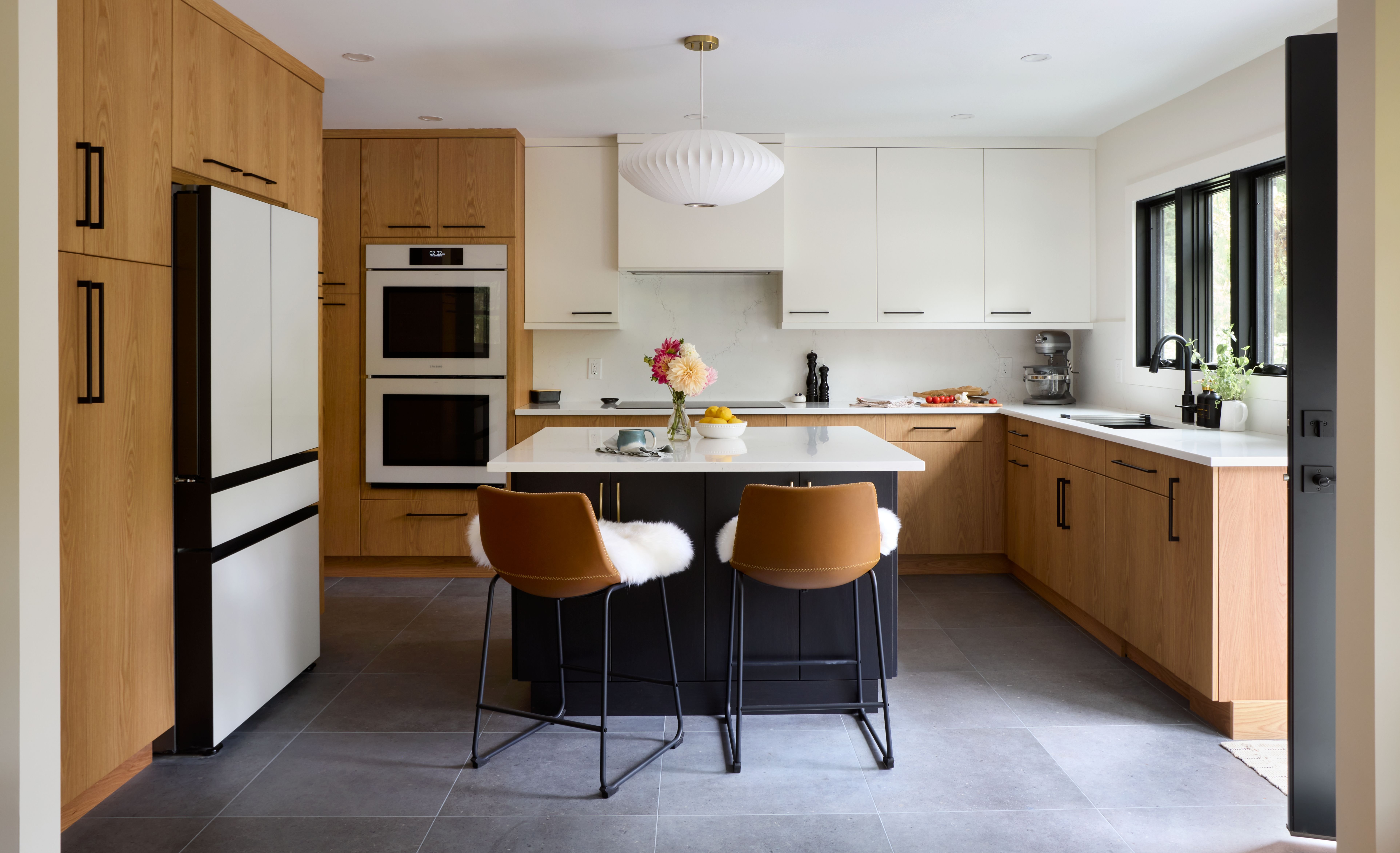 The image displays a modern kitchen with a combination of wood grain and white flat-panel cabinetry, a black island with a white countertop and two leather bar stools, a white refrigerator, double wall ovens, and dark gray tiled flooring.