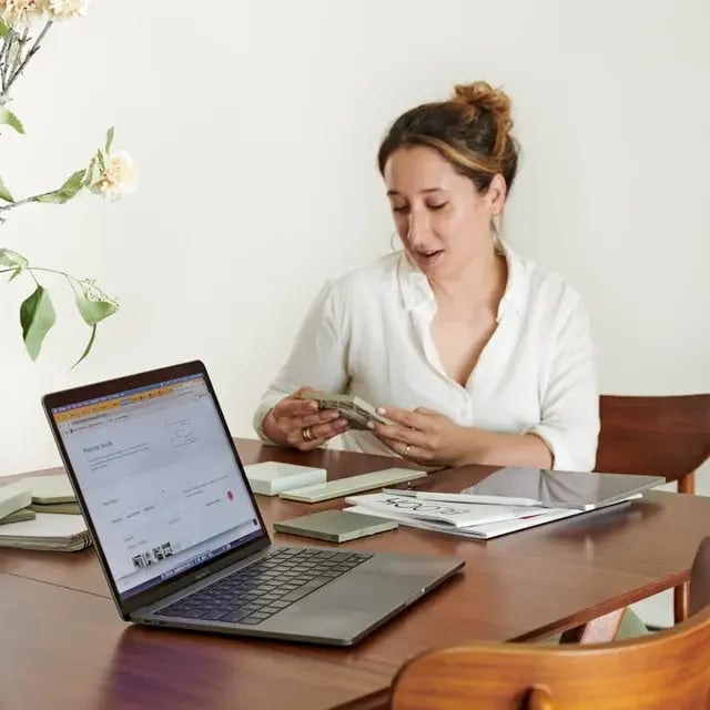 A design professional sitting at a table looking at tiles with an open laptop.