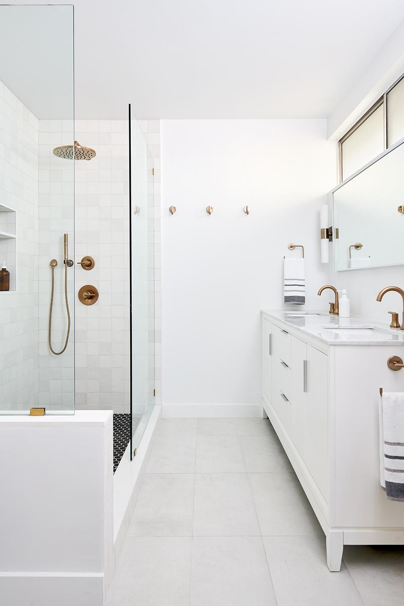 The image shows a modern, bright bathroom with white subway tile in the walk-in shower enclosure, which is separated by a glass partition, and a long white double vanity with gold-toned fixtures.