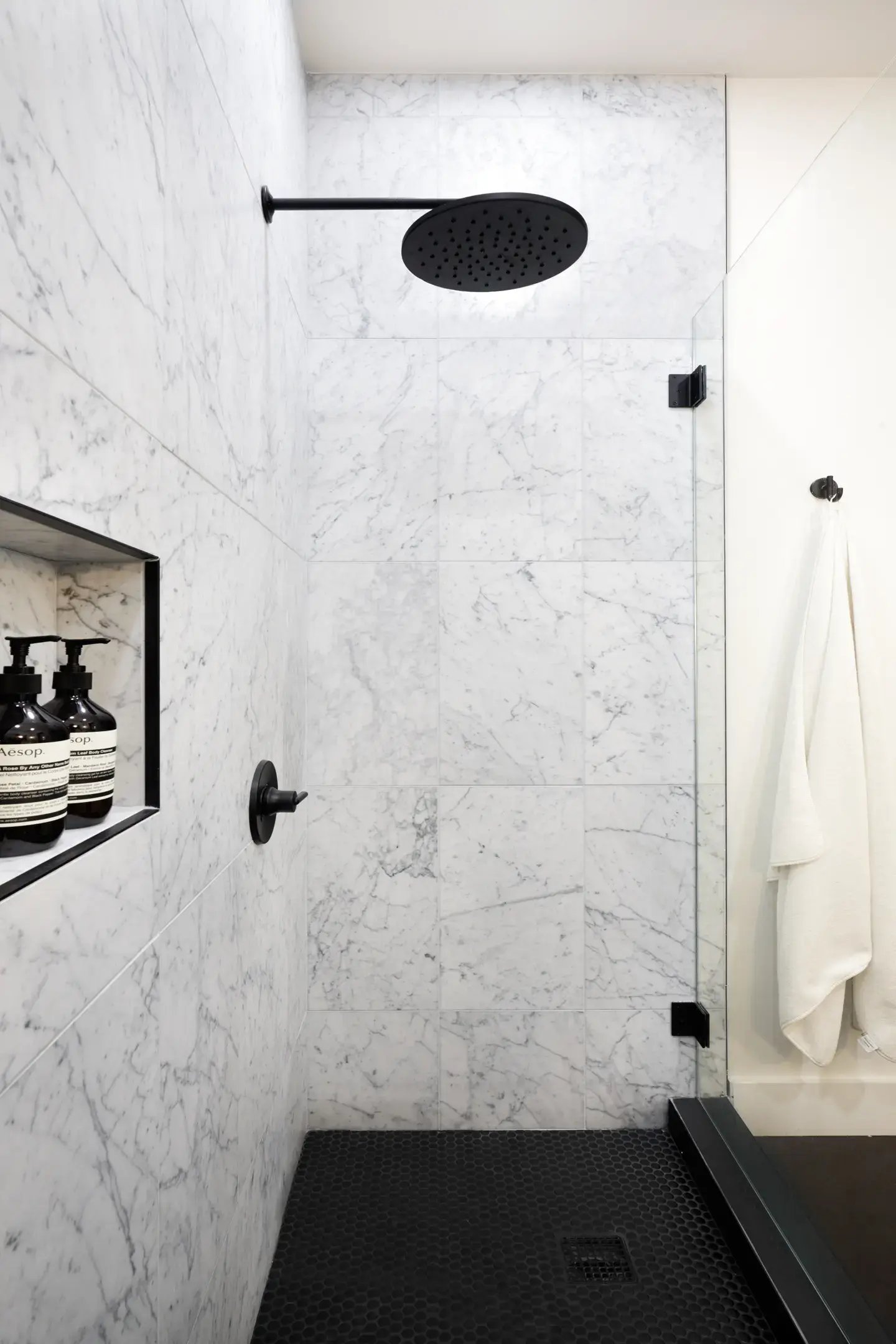 A minimalist shower with white marble wall tiles, matte black rain shower and fixtures, a recessed niche, clear glass panel, and dark hexagonal tile flooring for contrast.