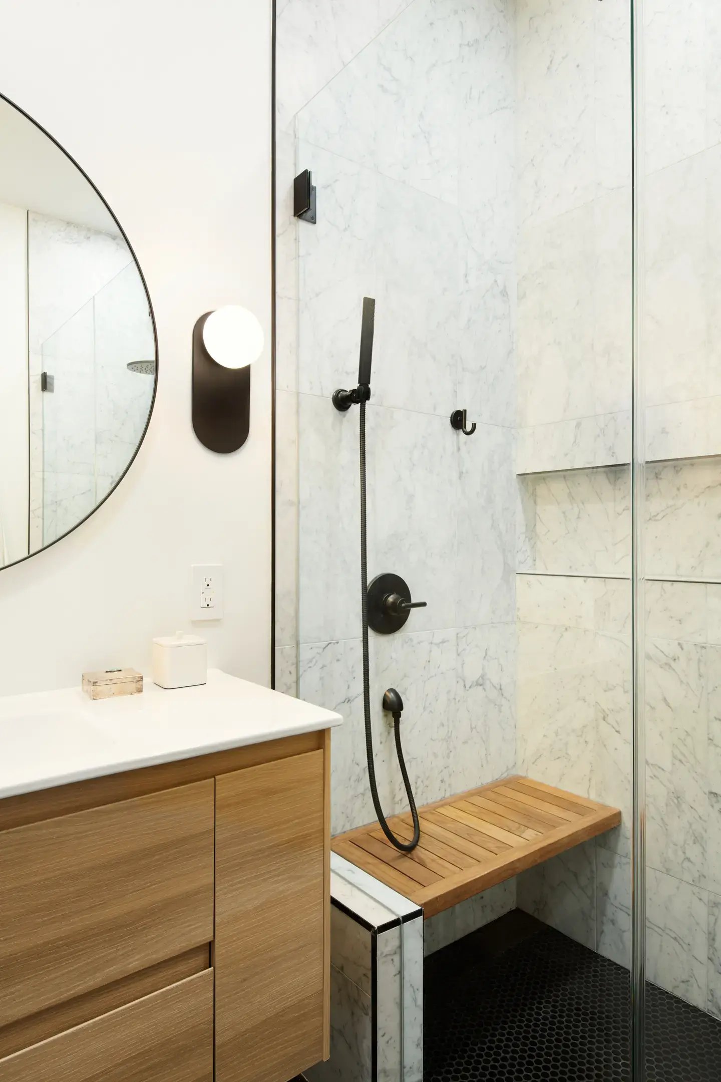 A modern bathroom features a light wood vanity with a white countertop, a circular mirror, a black wall sconce, and a walk-in shower with white marble-patterned tile, black hexagon floor tile, and a teak bench.