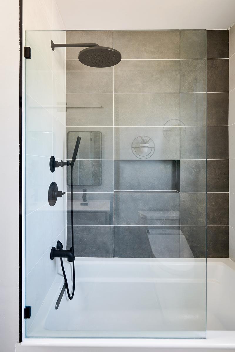 A modern bathroom features a glass shower enclosure with matte black fixtures, including a showerhead, a handheld sprayer, and a control set, against a tiled wall with a built-in niche, and a white bathtub is visible below.
