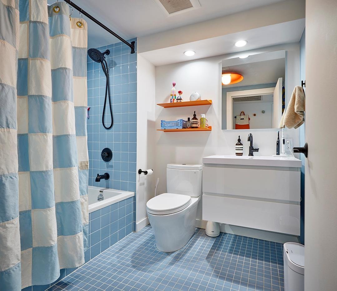 Compact bathroom with a blue tiled tub shower, matte black fixtures, patterned shower curtain, and a modern white vanity with open accent shelves.