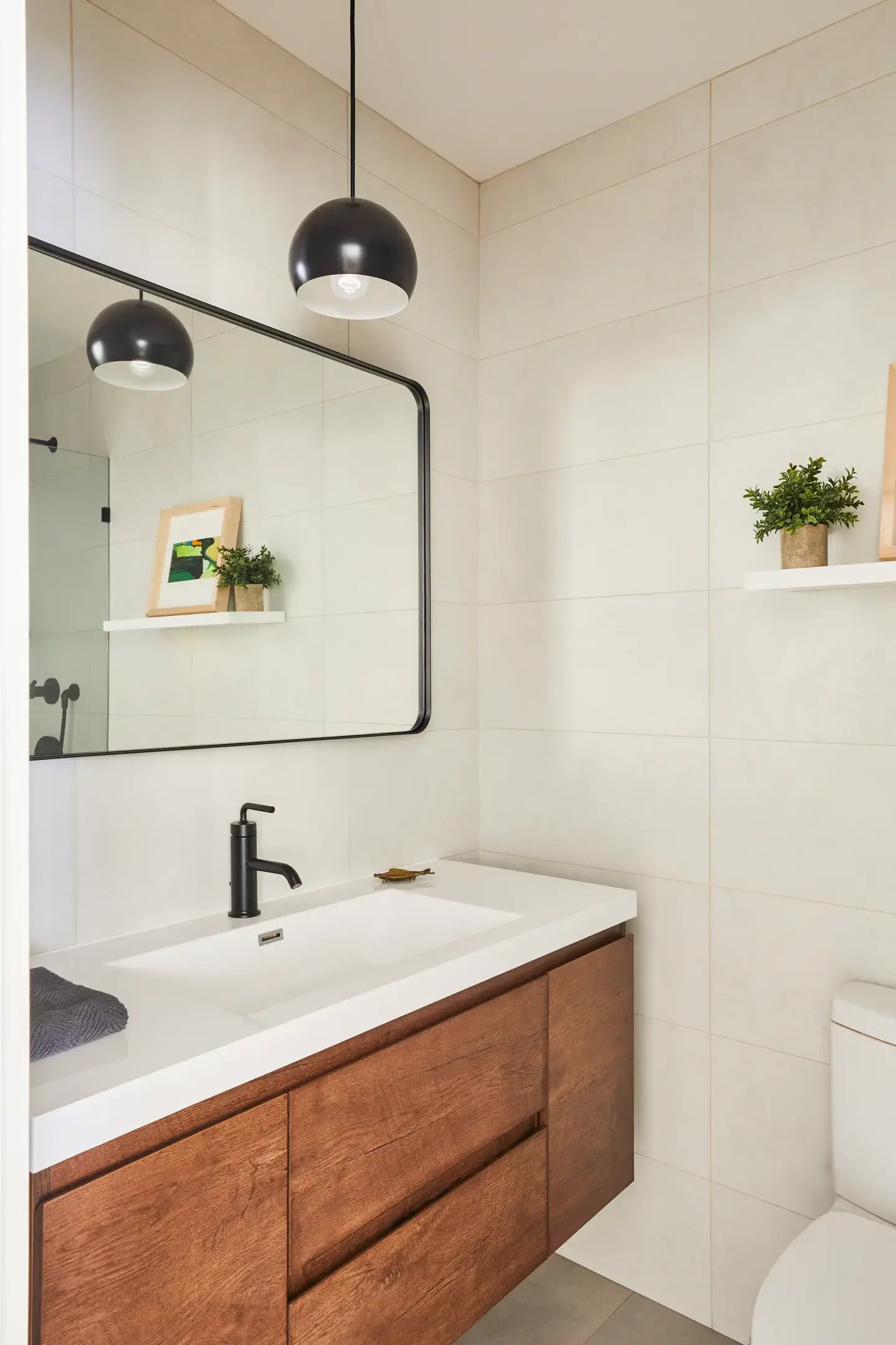 A modern, minimalist bathroom with a wooden vanity, white countertop and sink, black faucet, rectangular mirror, black pendant light, white tiled walls, and small decorative plants.