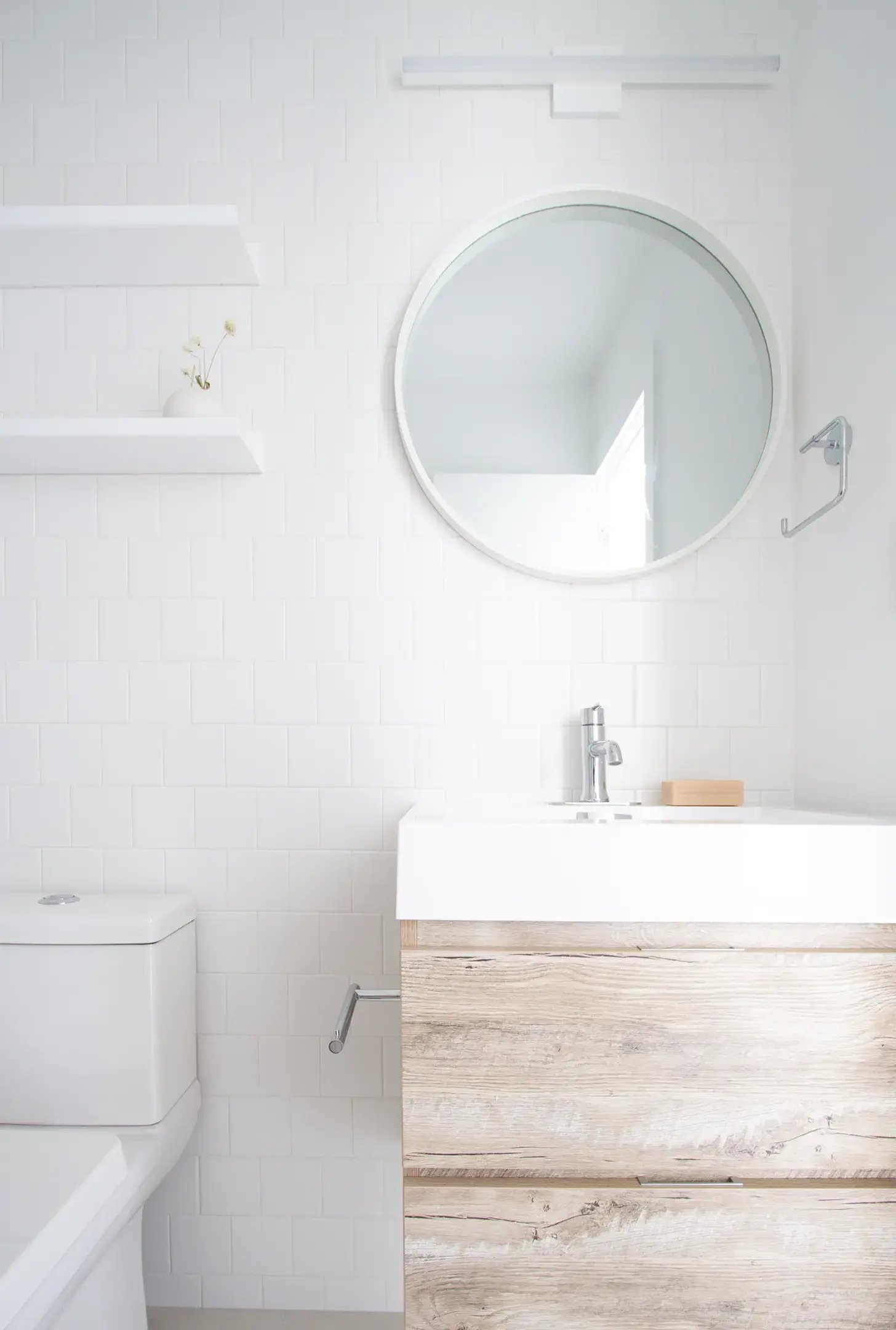 A minimalist white bathroom with round mirror and light wood vanity.