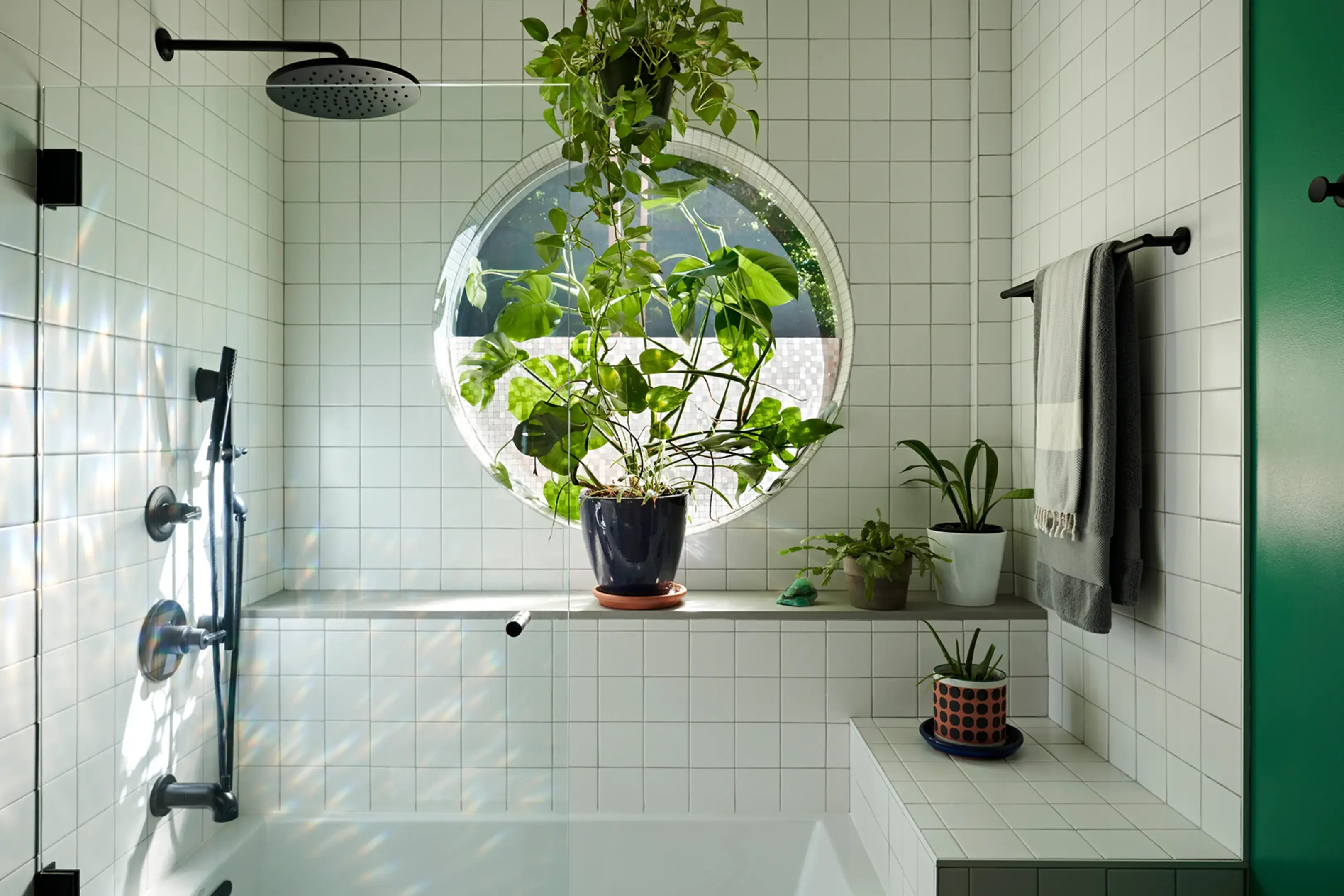 White tiled bathtub shower with matte black fixtures, a round window ledge filled with cascading green plants, and soft natural light creating a spa-like atmosphere.