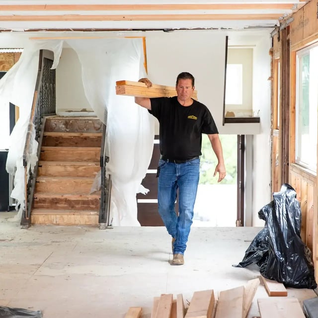 Construction worker carrying lumber inside a house under renovation.