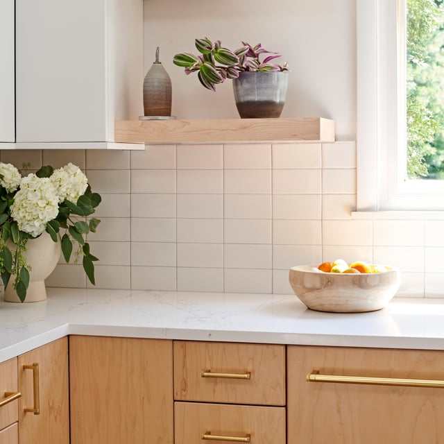 A modern kitchen featuring light wood cabinets with gold hardware, white countertops, and a white tiled backsplash. A wooden floating shelf holds decorative pottery and a plant.