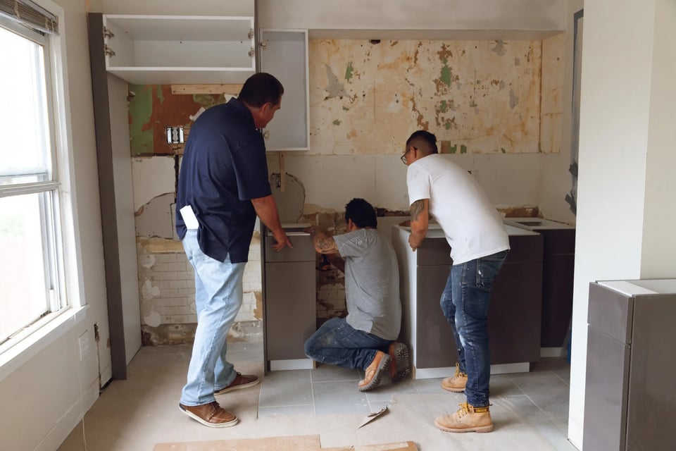 Three workers installing kitchen cabinets during a home renovation, with exposed walls and unfinished surfaces.