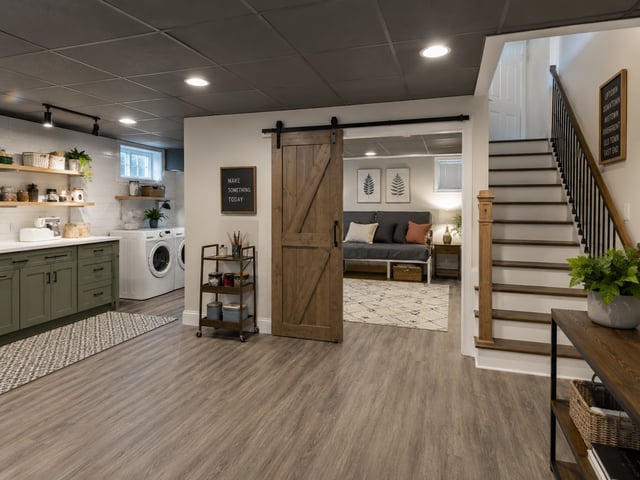 A modern semi-finished basement featuring a laundry area, wood-look flooring, and a sliding barn door leading to a lounge space.