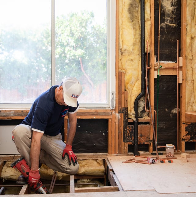 Worker kneels in gutted room installing plumbing and flooring