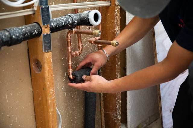 A worker installing foam insulation on copper pipes.