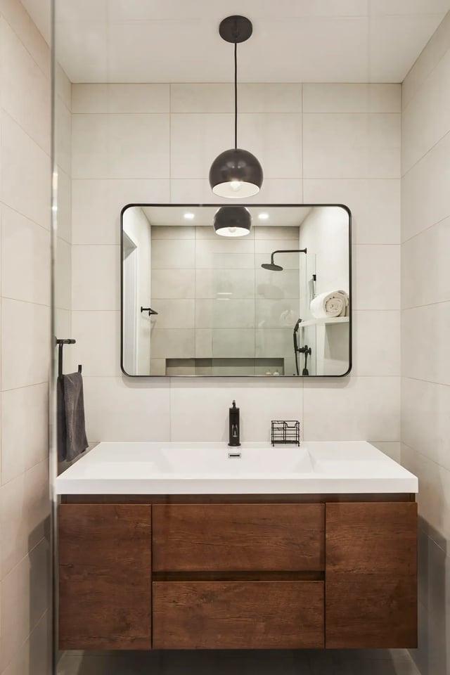 A modern bathroom featuring a floating wood vanity, a white countertop with a black faucet, and a large rectangular mirror with rounded corners.