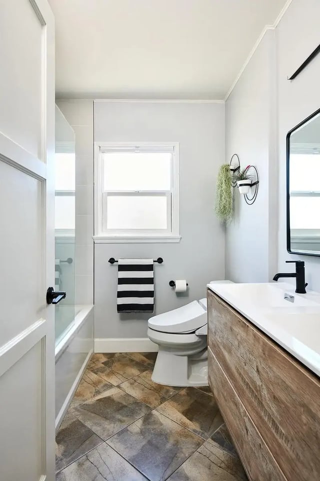 a modern bathroom with a white vanity featuring a wood-textured front, a white toilet, and a bathtub with a glass door. The room has grey walls, a small window, and diamond-patterned floor tiles.