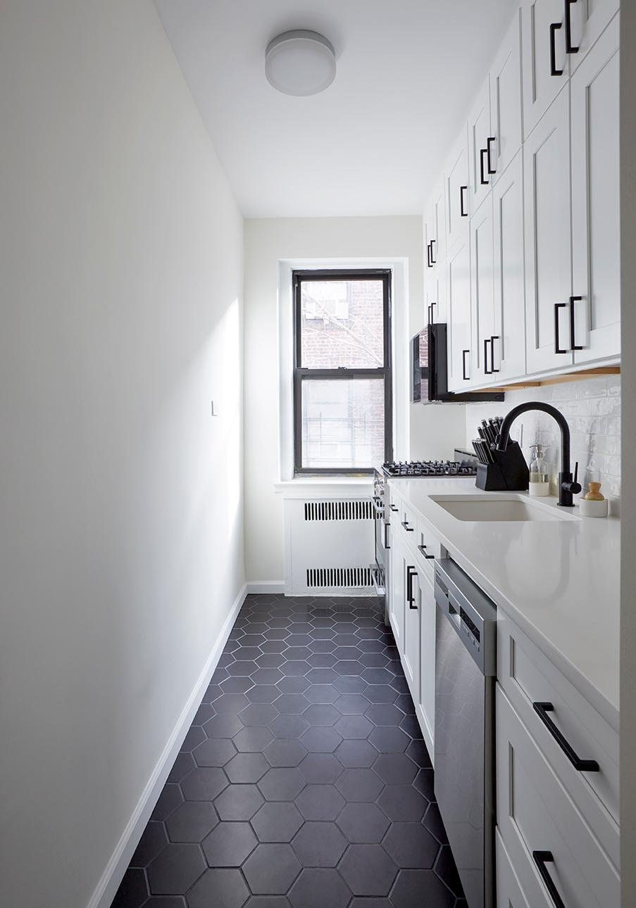 The image shows a narrow galley kitchen with white cabinetry and black hexagonal floor tiles.