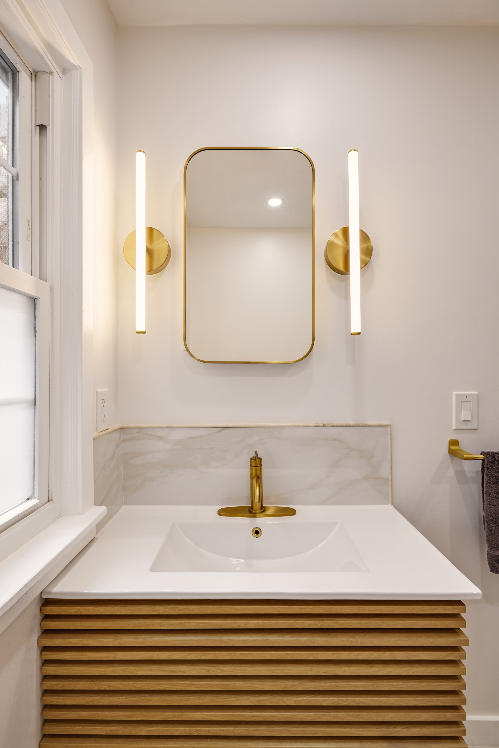 A modern bathroom features a slatted wood vanity cabinet with a white countertop and undermount sink, a brass single-hole faucet, and a rectangular, rounded-edge brass-framed mirror flanked by two vertical brass and glass tube vanity lights, with a white marble backsplash trimmed in brass and set against a light-colored wall.