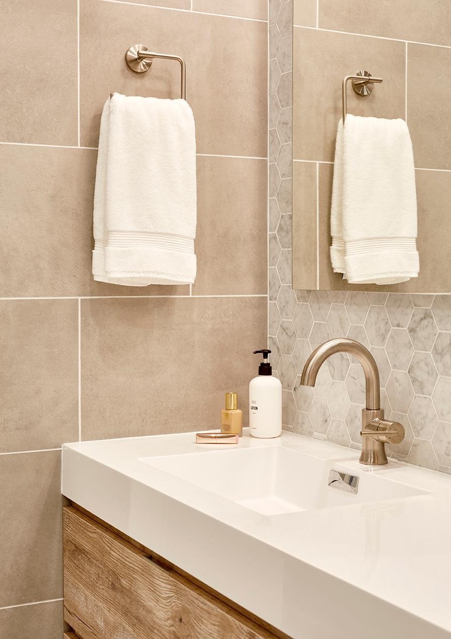 A white rectangular sink and brushed nickel faucet are set into a white countertop above a wooden vanity, with a white hand towel hanging on a brushed nickel bar against a wall of rectangular tan tiles and a small section of hexagonal marble tiles next to a mirror.