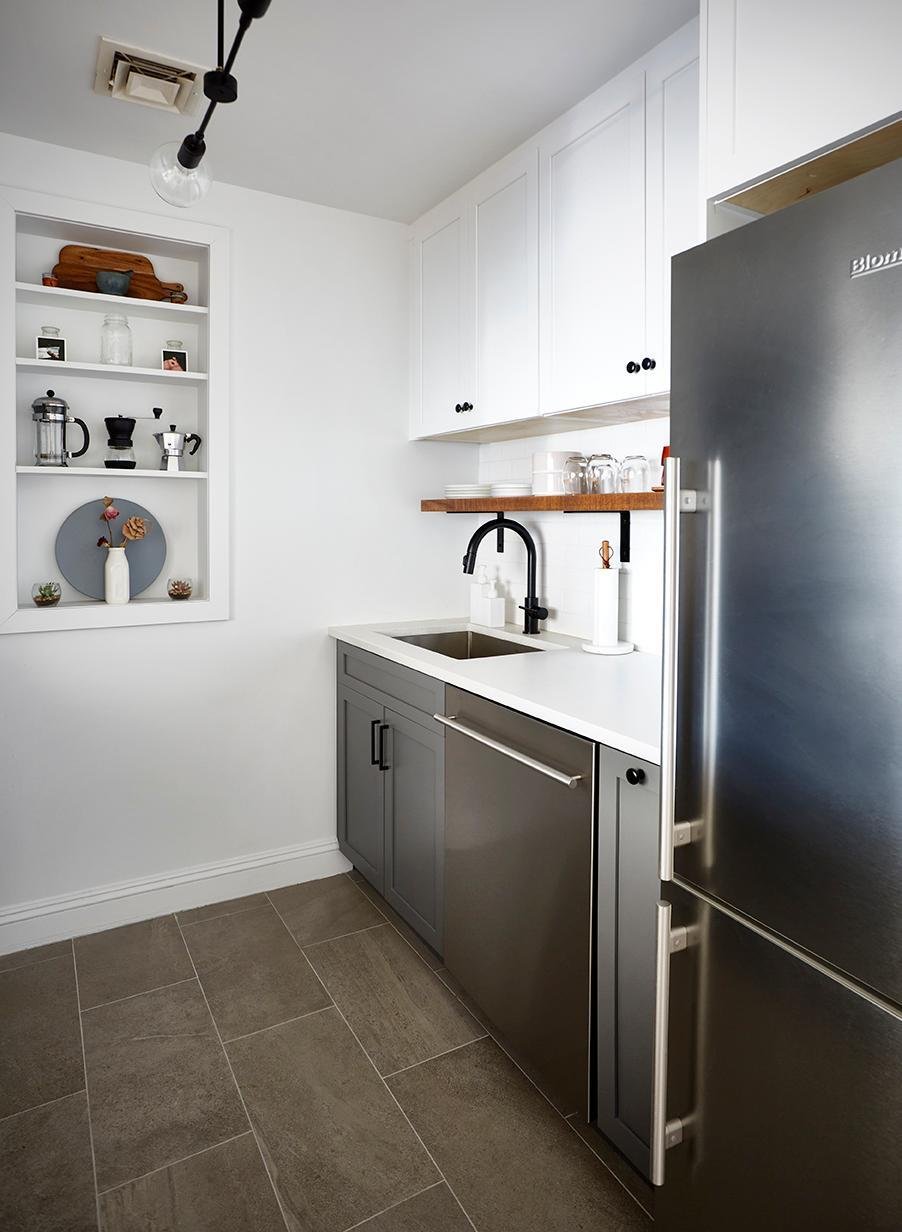 The image depicts a small, modern kitchen with gray base cabinets, white upper cabinets, a stainless steel refrigerator and dishwasher, a black gooseneck faucet, a white countertop, and a built-in recessed shelving unit.