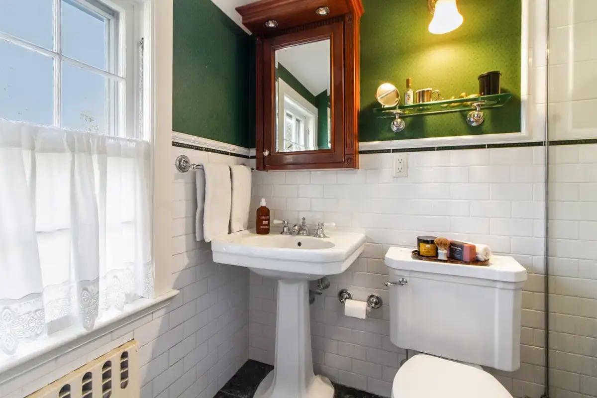 A traditionally styled bathroom featuring a pedestal sink, a wood medicine cabinet, and green walls above white subway tile.