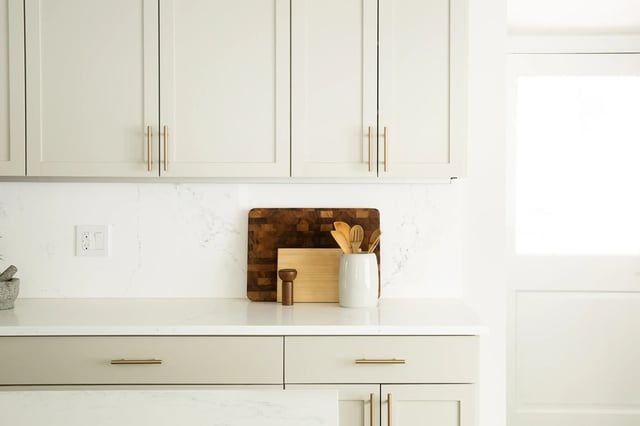 A modern kitchen features light beige upper and lower cabinets with gold hardware, a white marble backsplash, and a countertop displaying a wooden end-grain cutting board, a lighter wood cutting board, a salt and pepper grinder, and a white ceramic utensil holder filled with wooden spoons.