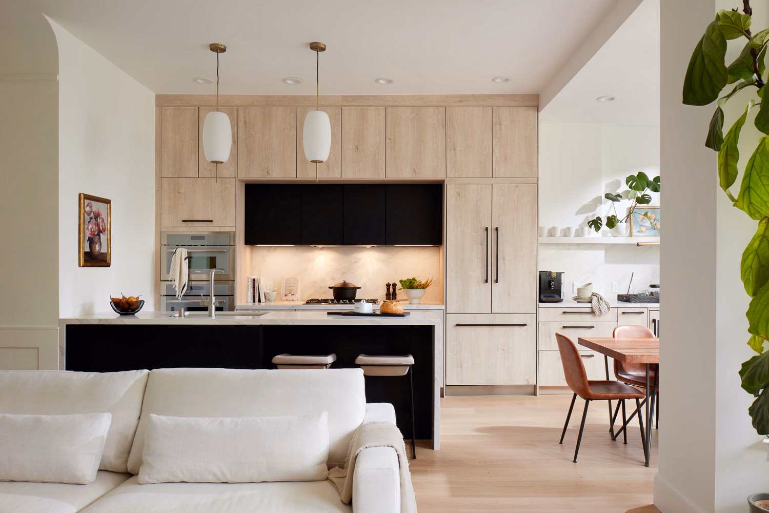  A sophisticated, open-concept kitchen and living area featuring sleek light-wood cabinetry, a bold black island with marble countertops, and elegant white pendant lighting.