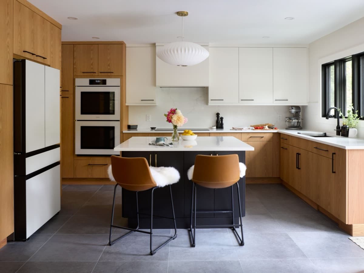 A modern kitchen with warm oak lower cabinets, white upper cabinets, a dark island with two leather stools and a gray tile floor